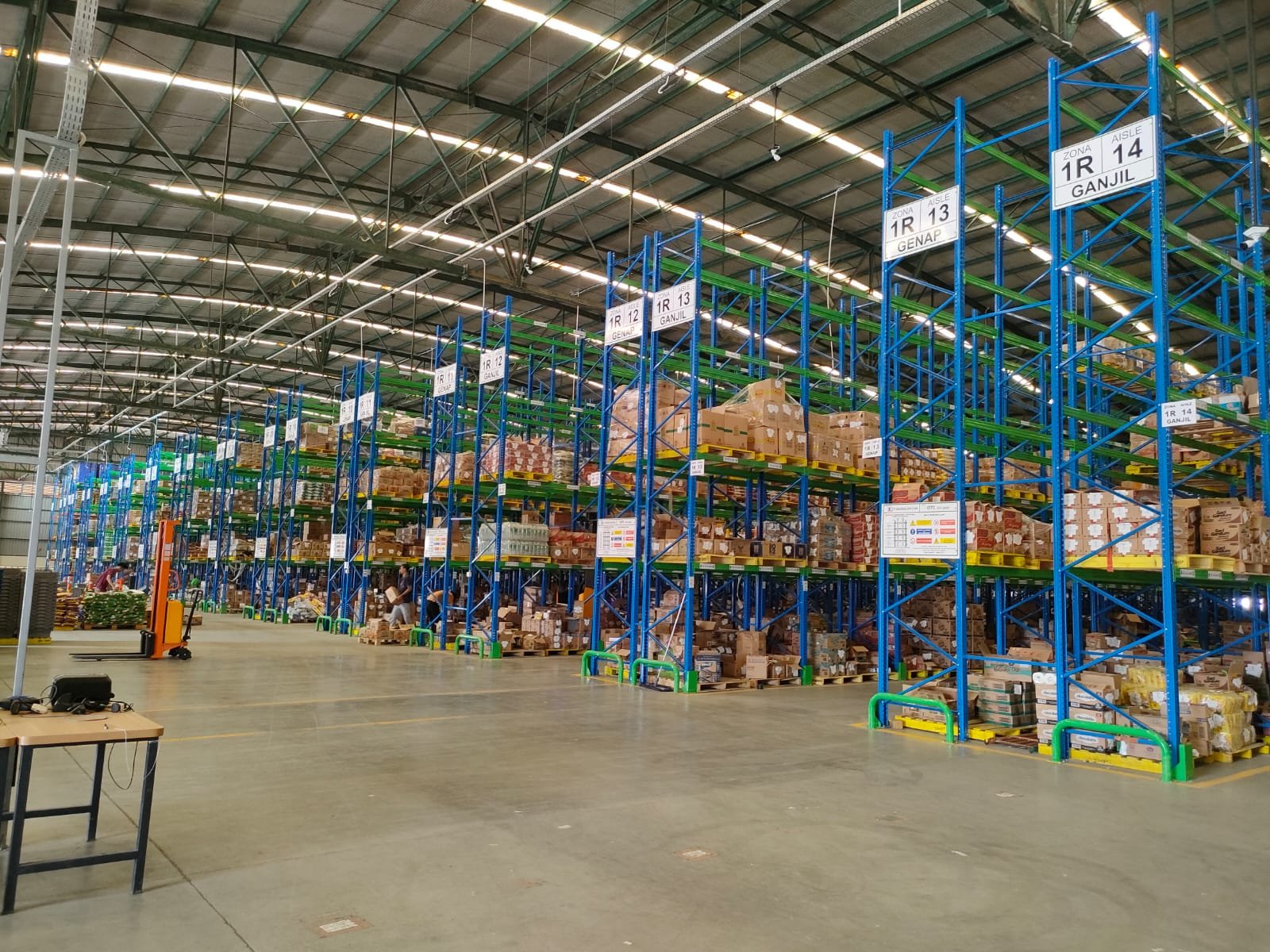 Rak gudang FMCG dengan pengaman kolom kuning dan label zona aisle
